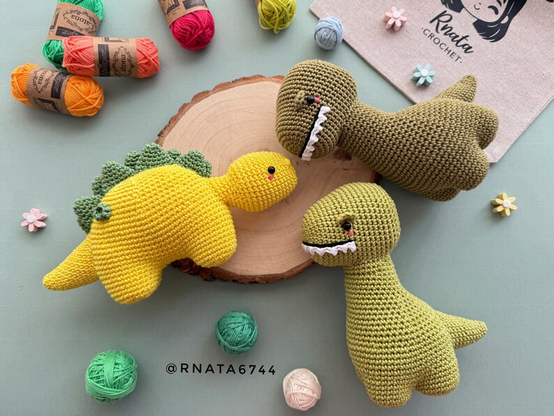May include: Crocheted dinosaur toys in yellow and green, with white teeth and red accents. The toys are displayed with colorful yarn balls and a wooden slice. The image includes the text "Rnata Crochet" and "@RNATA6744".