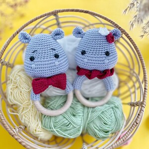 May include: Two crocheted hippo baby rattles in a wicker basket. The hippos are light blue with red bow ties and wooden rings. One hippo has a white flower on its head. The basket contains skeins of yellow and green yarn, set against a yellow background.