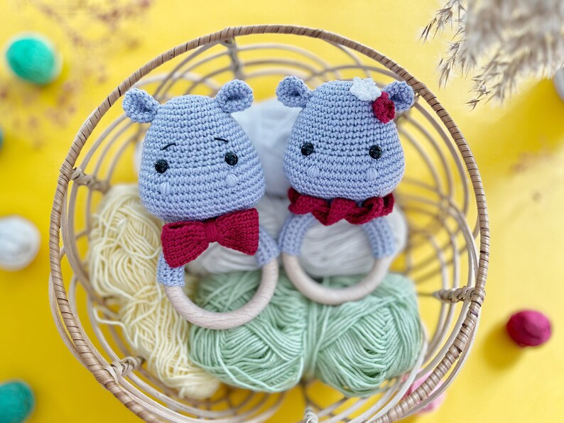 May include: Two crocheted hippo baby rattles in a wicker basket. The hippos are light blue with red bow ties and wooden rings. One hippo has a white flower on its head. The basket contains skeins of yellow and green yarn, set against a yellow background.