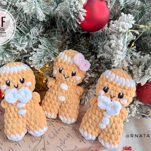 May include: Three handmade gingerbread man ornaments with white icing details, bows, and blush. The ornaments are set against a festive Christmas tree background with red ornaments and the text "PDF PATTERN ENGLISH".