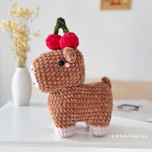 May include: A handmade brown crochet animal with a cherry on its head. The plush toy has a white muzzle, pink ears, and pink feet. The cherries are red with a green stem. The toy is on a white surface.