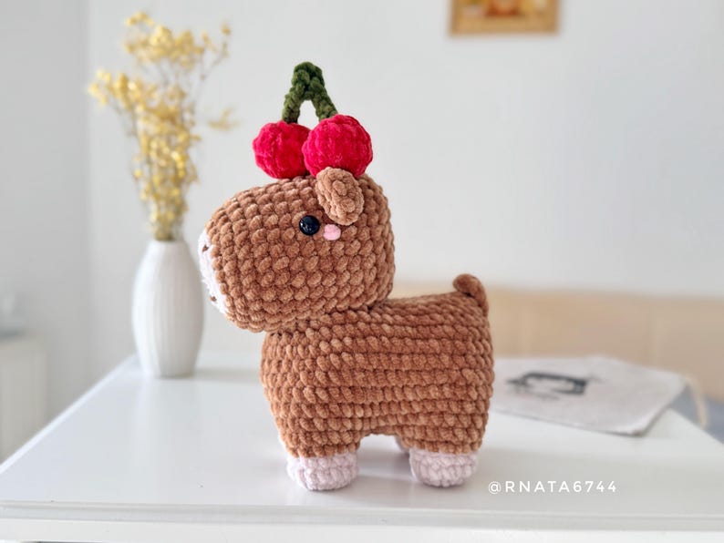 May include: A handmade brown crochet animal with a cherry on its head. The plush toy has a white muzzle, pink ears, and pink feet. The cherries are red with a green stem. The toy is on a white surface.