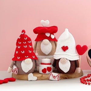 May include: Three crocheted gnomes wearing red and white hats with hearts. The gnomes are sitting on a wooden surface with a small red and white heart-shaped box and a red heart on a stick.