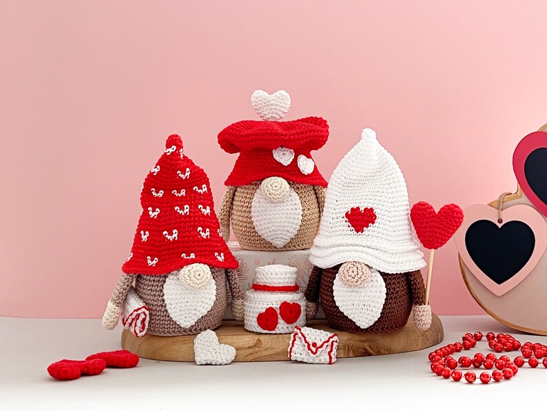 May include: Three crocheted gnomes wearing red and white hats with hearts. The gnomes are sitting on a wooden surface with a small red and white heart-shaped box and a red heart on a stick.