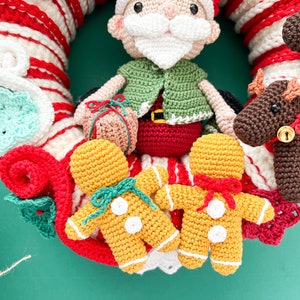 May include: A crocheted Christmas wreath with a Santa Claus, two gingerbread men, and a reindeer. The wreath is decorated with red and white stripes and green and white crocheted flowers.