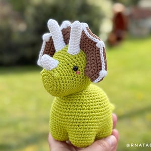 May include: A handmade, crocheted triceratops dinosaur in shades of green, brown, and white. The dinosaur has three horns, a frill, and a friendly expression. The toy is shown against a blurred green background.