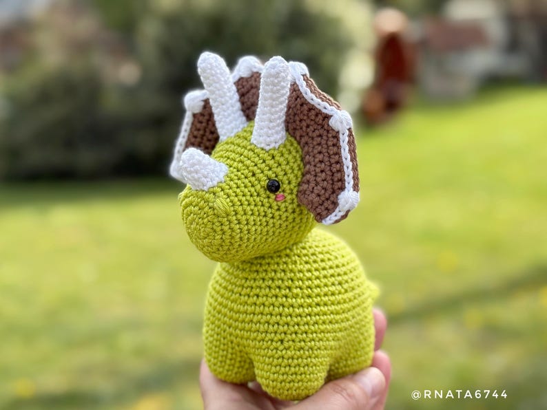 May include: A handmade, crocheted triceratops dinosaur in shades of green, brown, and white. The dinosaur has three horns, a frill, and a friendly expression. The toy is shown against a blurred green background.