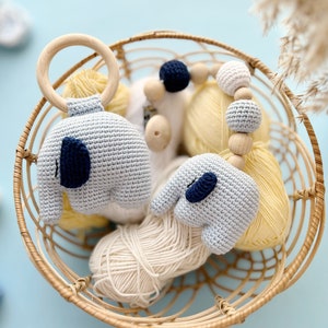 Puede incluir: Una cesta de mimbre llena de hilo, un anillo de madera con un elefante de crochet unido y un collar de cuentas de crochet en forma de elefante. El hilo es en tonos crema y amarillo, y los elefantes son azul claro con detalles en azul marino.
