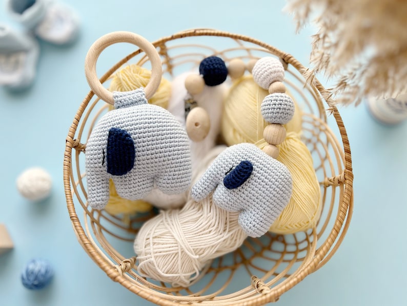 Puede incluir: Una cesta de mimbre llena de hilo, un anillo de madera con un elefante de crochet unido y un collar de cuentas de crochet en forma de elefante. El hilo es en tonos crema y amarillo, y los elefantes son azul claro con detalles en azul marino.