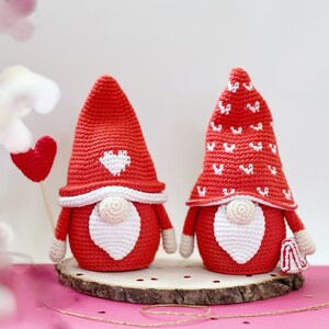 May include: Two crocheted gnomes with red hats and white beards. One gnome has a white heart on its hat and the other has a white pattern of hearts on its hat. The gnomes are sitting on a wooden base.