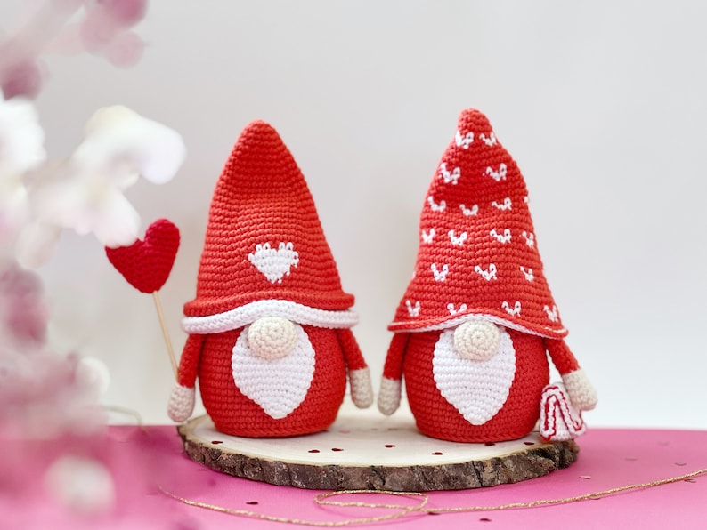 May include: Two crocheted gnomes with red hats and white beards. One gnome has a white heart on its hat and the other has a white pattern of hearts on its hat. The gnomes are sitting on a wooden base.