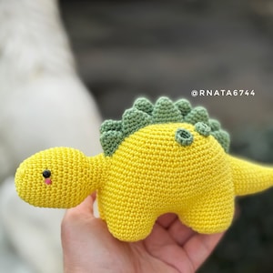 May include: A handmade crochet dinosaur toy in bright yellow with green accents. The dinosaur has a small head, a long tail, and a row of green spikes down its back. The toy is soft and appears to be made of yarn.