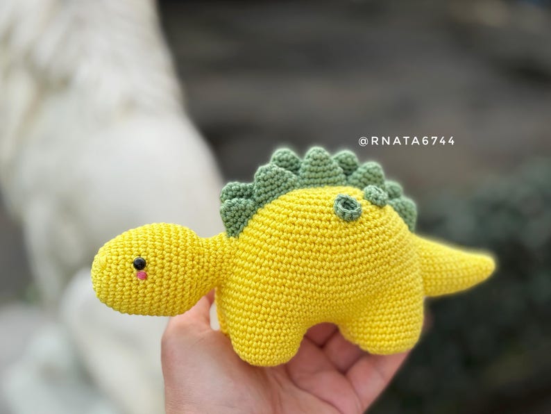 May include: A handmade crochet dinosaur toy in bright yellow with green accents. The dinosaur has a small head, a long tail, and a row of green spikes down its back. The toy is soft and appears to be made of yarn.