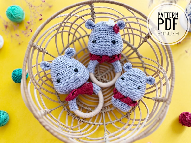 May include: Three crocheted hippo baby rattles in a woven basket. The hippos are light blue with red bow ties and wooden rings. The image includes the text "PATTERN PDF ENGLISH".