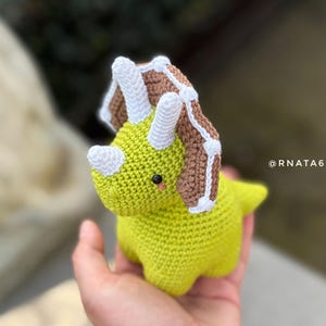 May include: A handmade, crocheted triceratops dinosaur in shades of green, brown, and white. The dinosaur has a friendly expression with small black eyes and a pink blush. The text "@RNATA6744" is visible.