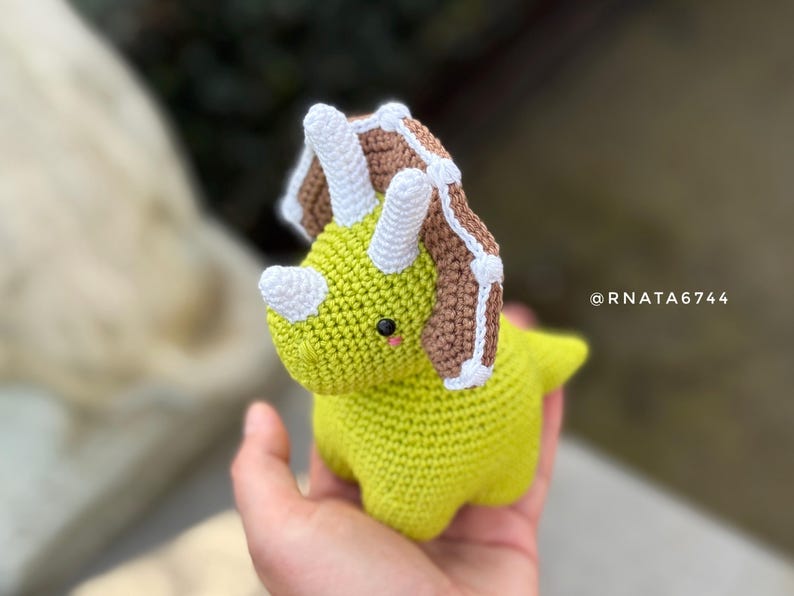 May include: A handmade, crocheted triceratops dinosaur in shades of green, brown, and white. The dinosaur has a friendly expression with small black eyes and a pink blush. The text "@RNATA6744" is visible.