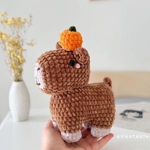 May include: A handmade brown crochet animal with a small orange pumpkin on its head. The plush toy has pink accents on its face and feet. The background includes a white vase with yellow flowers.