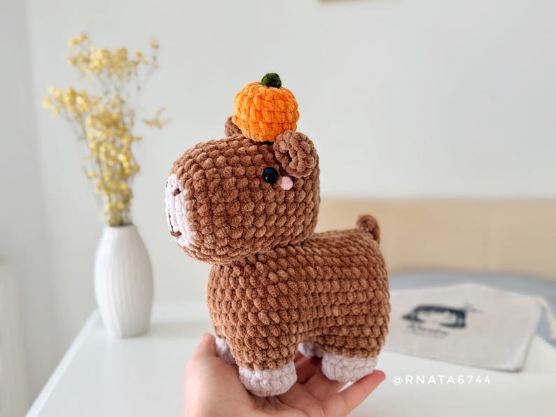 May include: A handmade brown crochet animal with a small orange pumpkin on its head. The plush toy has pink accents on its face and feet. The background includes a white vase with yellow flowers.