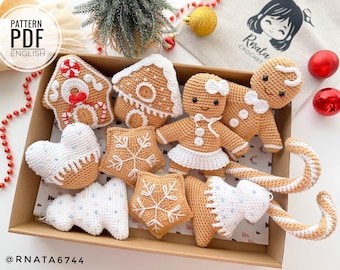 May include: A collection of crocheted gingerbread ornaments in a box. The ornaments include a gingerbread house, gingerbread people, a gingerbread star, and a gingerbread candy cane. The ornaments are all brown and white and have a festive Christmas theme.