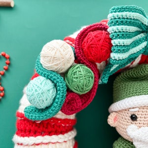 May include: A crocheted Christmas wreath with red, white, and green yarn. The wreath is decorated with a green and white striped bow and small balls of yarn in various colors. A crocheted Santa Claus ornament is attached to the wreath.