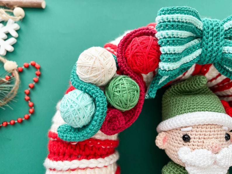 May include: A crocheted Christmas wreath with red, white, and green yarn. The wreath is decorated with a green and white striped bow and small balls of yarn in various colors. A crocheted Santa Claus ornament is attached to the wreath.