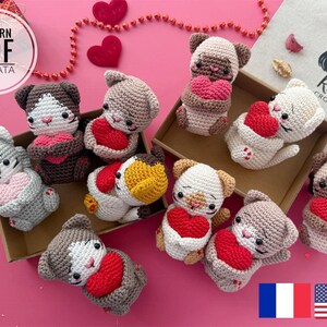 May include: A set of nine crocheted cat amigurumi toys holding red hearts. The cats are in various colors, including gray, white, and brown. The toys are arranged in a box with a pink background. The text "Rnata Crochet" is visible on the box.