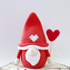 May include: A red crocheted gnome with a white heart on its hat and a red heart balloon. The gnome is sitting on a wooden slice with red glitter hearts scattered around it.
