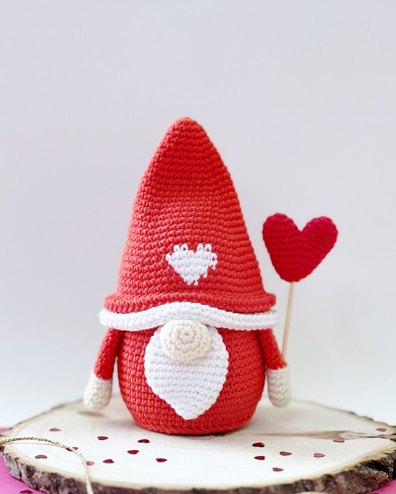 May include: A red crocheted gnome with a white heart on its hat and a red heart balloon. The gnome is sitting on a wooden slice with red glitter hearts scattered around it.