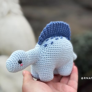 May include: A light blue crocheted dinosaur toy with a dark blue crest and dotted details. The handmade plushie has a long neck, small legs, and a cute face with a pink blush. The toy is approximately 5 inches long.