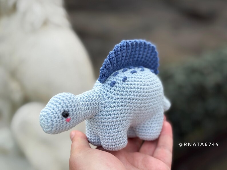 May include: A light blue crocheted dinosaur toy with a dark blue crest and dotted details. The handmade plushie has a long neck, small legs, and a cute face with a pink blush. The toy is approximately 5 inches long.