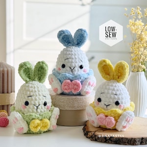 Może przedstawiać: Trzy ręcznie robione figurki króliczków na szydełku w kolorze białym, z niebieskimi, zielonymi i żółtymi akcentami. Każdy króliczek ma oklapnięte uszy, kokardę i różowe podkładki. Widoczny jest tekst "LOW-SEW PATTERN".