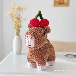 May include: A handmade brown crochet animal with a light pink face and feet, topped with two red cherries and a green stem. The plush toy is on a white surface.