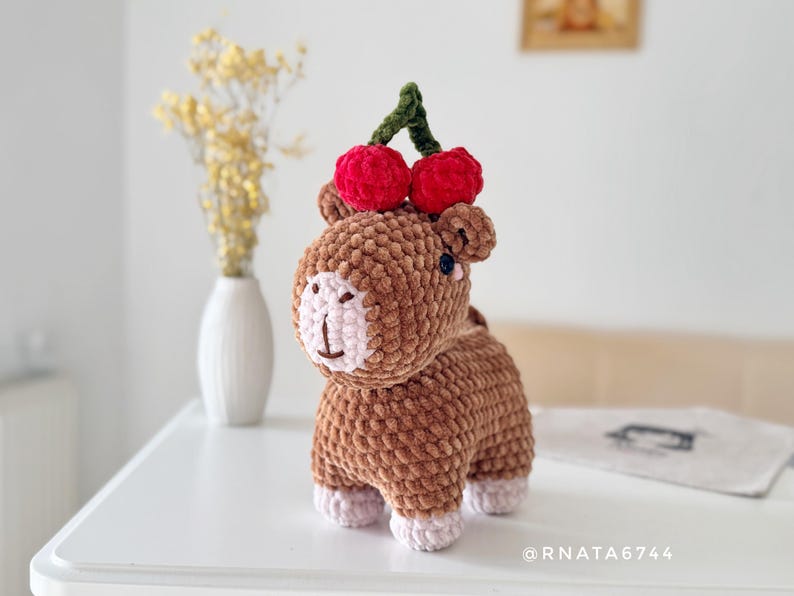 May include: A handmade brown crochet animal with a light pink face and feet, topped with two red cherries and a green stem. The plush toy is on a white surface.