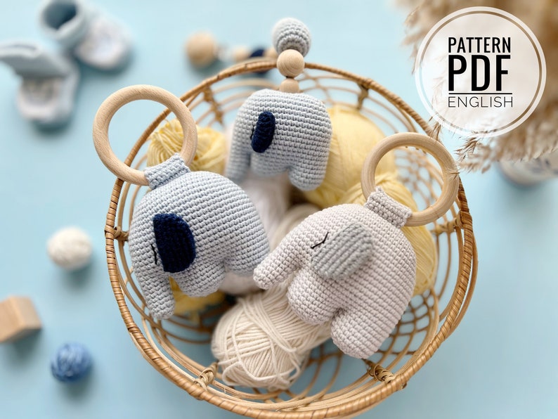 Puede incluir: Tres juguetes de elefante de crochet en una cesta de mimbre. Dos de los elefantes son de color azul claro y uno es de color gris claro. Los elefantes est&aacute;n unidos a anillos de madera. La cesta est&aacute; llena de ovillos de lana.