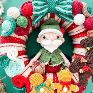 May include: A crocheted Christmas wreath with a red and white candy cane stripe design. The wreath is decorated with a green and white bow, a crocheted elf, gingerbread people, and reindeer.