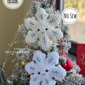 Könnte beinhalten: Zwei weiße, plüschige Schneeflocken-Ornamente mit blauen Blattdetails und lächelnden Gesichtern werden auf einem Weihnachtsbaum gezeigt. Die Ornamente sind handgefertigt und tragen ein "NO SEW"-Etikett. Das Bild enthält auch ein "PDF PATTERN ENGLISH ONLY"-Etikett.