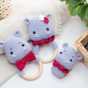 May include: Three handmade crochet hippo baby rattles. Each rattle features a grey hippo head with black eyes and a red bow or collar. The rattles have wooden ring handles. One hippo has a white flower.