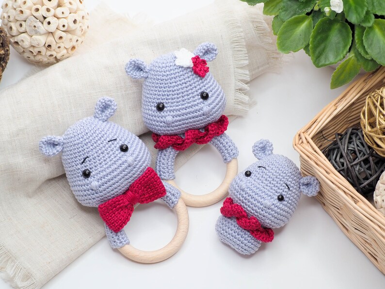 May include: Three handmade crochet hippo baby rattles. Each rattle features a grey hippo head with black eyes and a red bow or collar. The rattles have wooden ring handles. One hippo has a white flower.