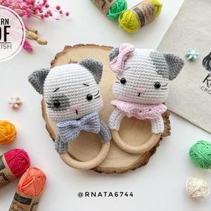 Può includere: Due giocattoli a sonagli a forma di gatto all'uncinetto con anelli di legno. Un gatto indossa un fiocco rosa e l'altro un fiocco blu. I giocattoli sono su una superficie di legno con gomitoli di lana e fiori sullo sfondo. Il testo "RNATA CROCHET" è visibile su una borsa di tela bianca.