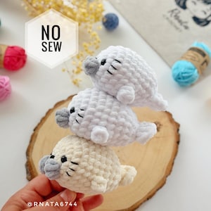 Puede incluir: Tres focas de ganchillo apiladas hechas de hilo suave blanco y crema con hocicos grises y ojos negros. La foca superior tiene una superposición hexagonal blanca con el texto NO SEW. Los peluches presentan un aspecto de punto grueso y texturizado.