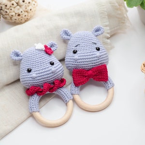 May include: Two crocheted hippo baby rattles. One has a red bow tie, the other a red ruffle and a white flower. Both have wooden rings and are light grey with black eyes.