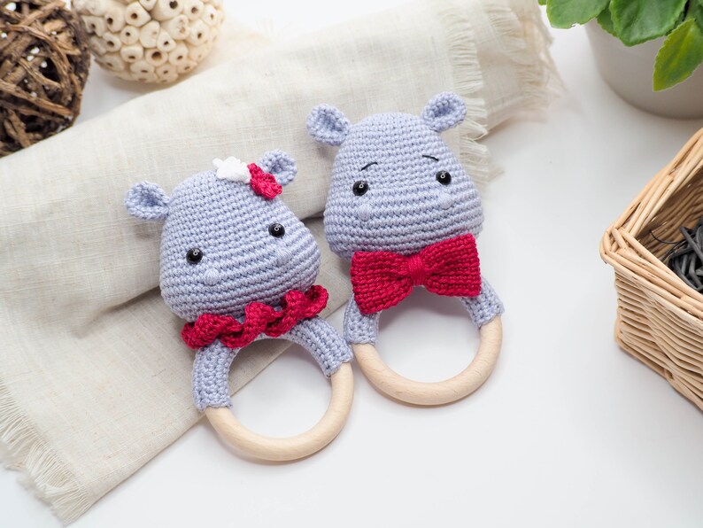 May include: Two crocheted hippo baby rattles. One has a red bow tie, the other a red ruffle and a white flower. Both have wooden rings and are light grey with black eyes.