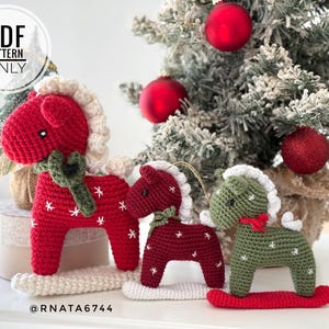 May include: Three crocheted rocking horse ornaments in red and olive green with white manes, tails, and embroidered snowflake details. The horses feature small black eyes and are mounted on white or red rockers. Text reads PDF PATTERN ONLY @RNATA6744.