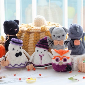 Puede incluir: Una colección de juguetes de ganchillo con temática de Halloween, que incluye un gato negro, un fantasma, una calabaza, un búho y un murciélago. Los juguetes están dispuestos sobre una superficie blanca con hojas y bayas de color otoñal.