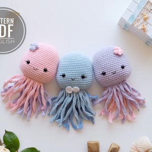 May include: Three crocheted jellyfish toys in pink, blue, and purple. Each jellyfish has a smiling face with black eyes and a small flower on top. The jellyfish are on a white surface.