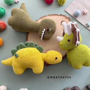 May include: Crocheted dinosaur toys in shades of yellow, green, and brown. The toys include a triceratops, a stegosaurus, and a T-Rex. Yarn and a wooden slice are also in the image. The artist's name, Rnata Crochet, is visible.