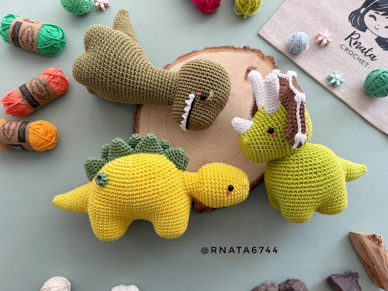 May include: Crocheted dinosaur toys in shades of yellow, green, and brown. The toys include a triceratops, a stegosaurus, and a T-Rex. Yarn and a wooden slice are also in the image. The artist's name, Rnata Crochet, is visible.