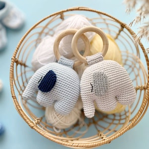 Puede incluir: Dos juguetes de elefante de crochet con detalles azules y grises unidos a anillos de madera. Los juguetes est&aacute;n en una cesta de mimbre con ovillos de lana.