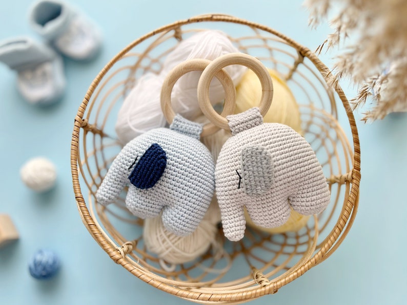 Puede incluir: Dos juguetes de elefante de crochet con detalles azules y grises unidos a anillos de madera. Los juguetes est&aacute;n en una cesta de mimbre con ovillos de lana.