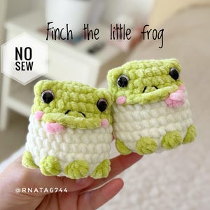 May include: Two handmade crochet frog toys. The frogs are light green and white with pink accents. The text "Finch the little frog" is above the frogs. The text "NO SEW" is in a white hexagon. The frogs have black eyes.